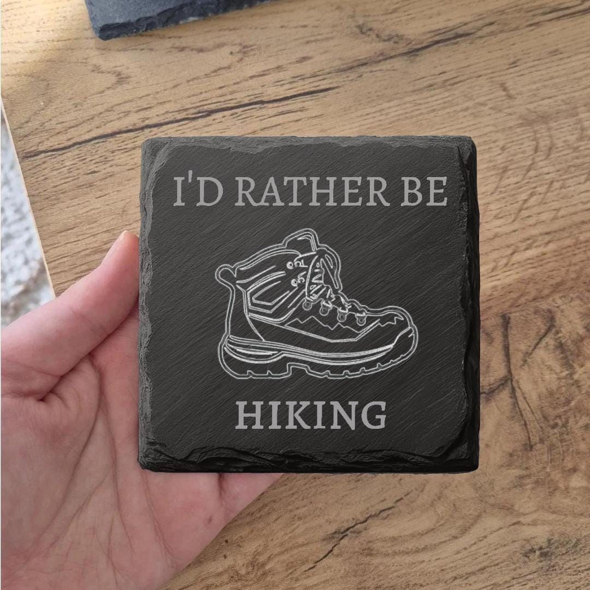 A hand holding a slate coaster with the text "I'D RATHER BE HIKING" and an illustration of a hiking boot.