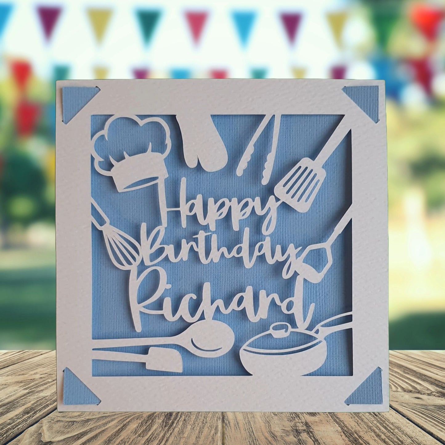A personalised birthday card for a chef, featuring a blue background with images of kitchen utensils and a chef's toque, with the name printed on the front. The card has a blue background.