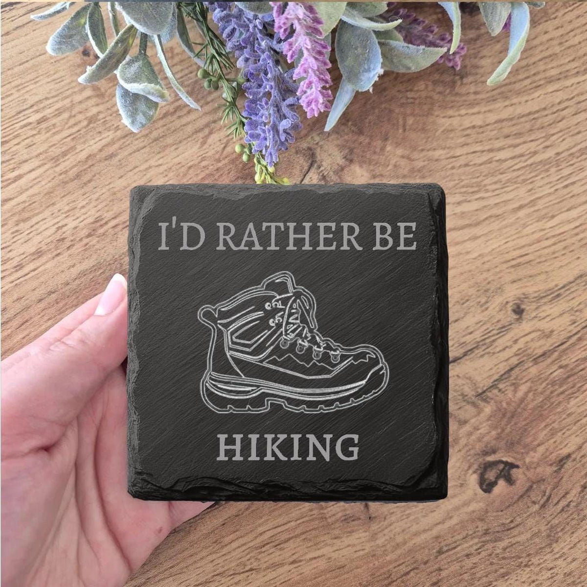 A hand holding a slate with a graphic of a hiking boot and the text "I'D RATHER BE HIKING" printed on it.