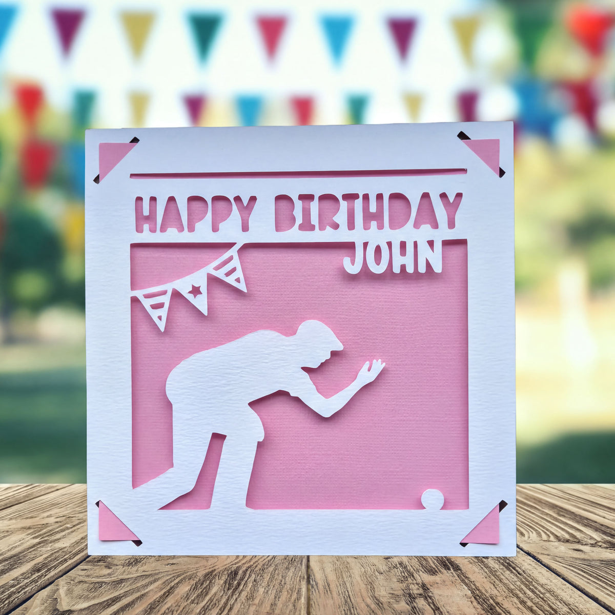 A white greeting card with a silhouette of a person kneeling, playing crown green bowling and a banner with a star, placed on a wooden surface.