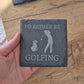 A hand holding a slate coaster with the text "I'D RATHER BE GOLFING" printed on it.