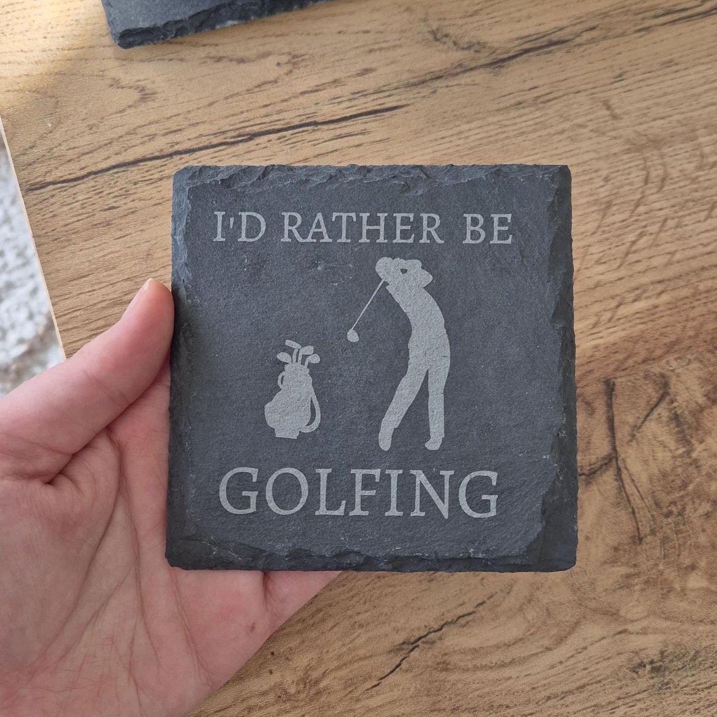 A hand holding a slate coaster with the text "I'D RATHER BE GOLFING" printed on it.