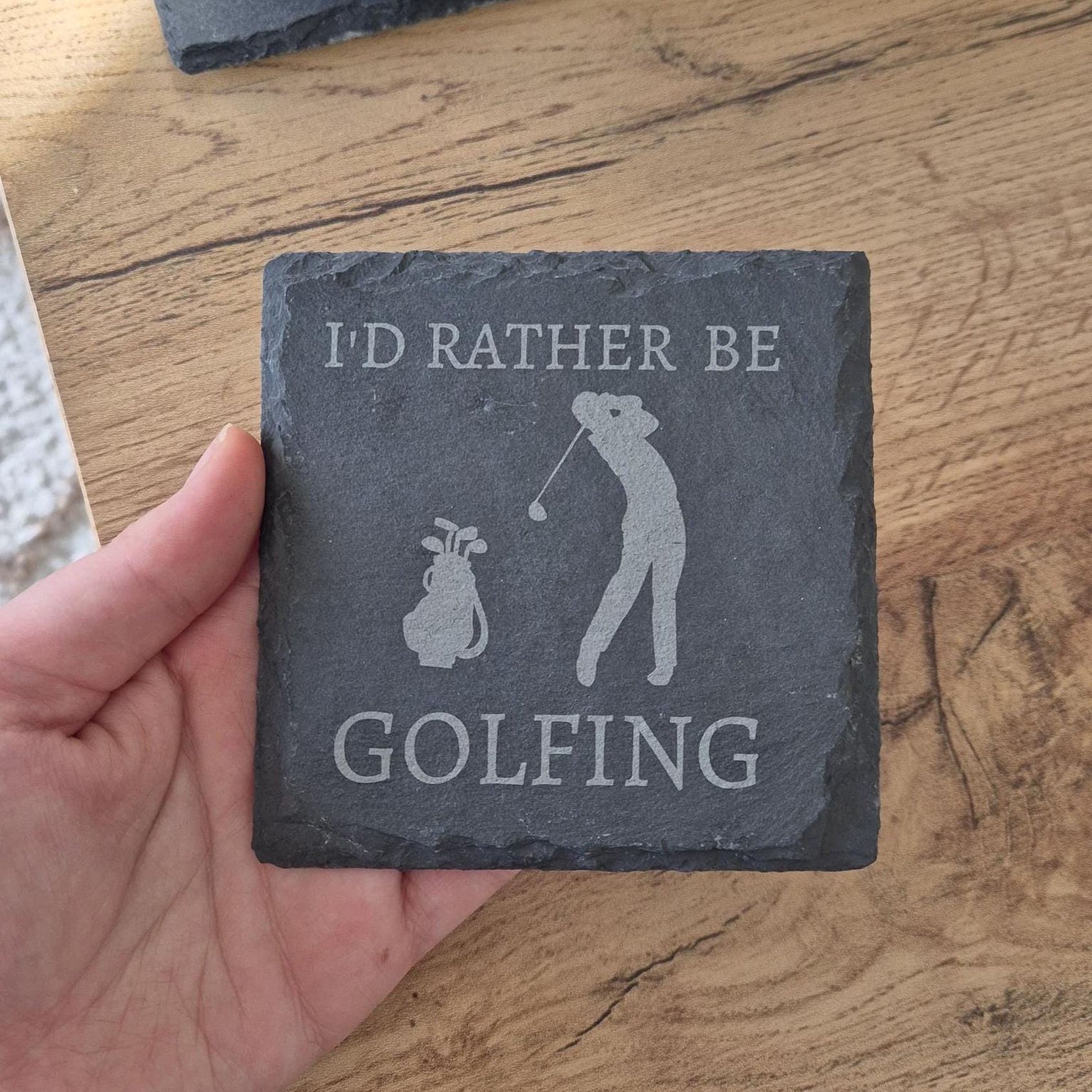 A hand holding a slate coaster with the text "I'D RATHER BE GOLFING" printed on it.