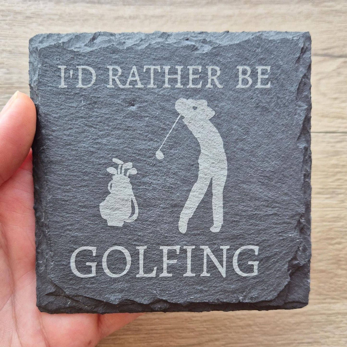 A hand holding a slate coaster with the text "I'd rather be GOLFING" and an illustration of a golfer.