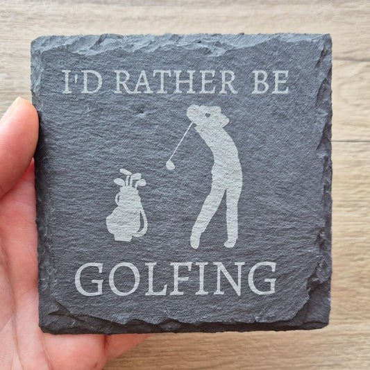 A hand holding a slate coaster with the text "I'd rather be GOLFING" and an illustration of a golfer.