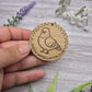 Wooden round keychain with pigeon illustration and 'Pigeon Appreciation Club' text, held by a hand on a wooden surface with flowers.