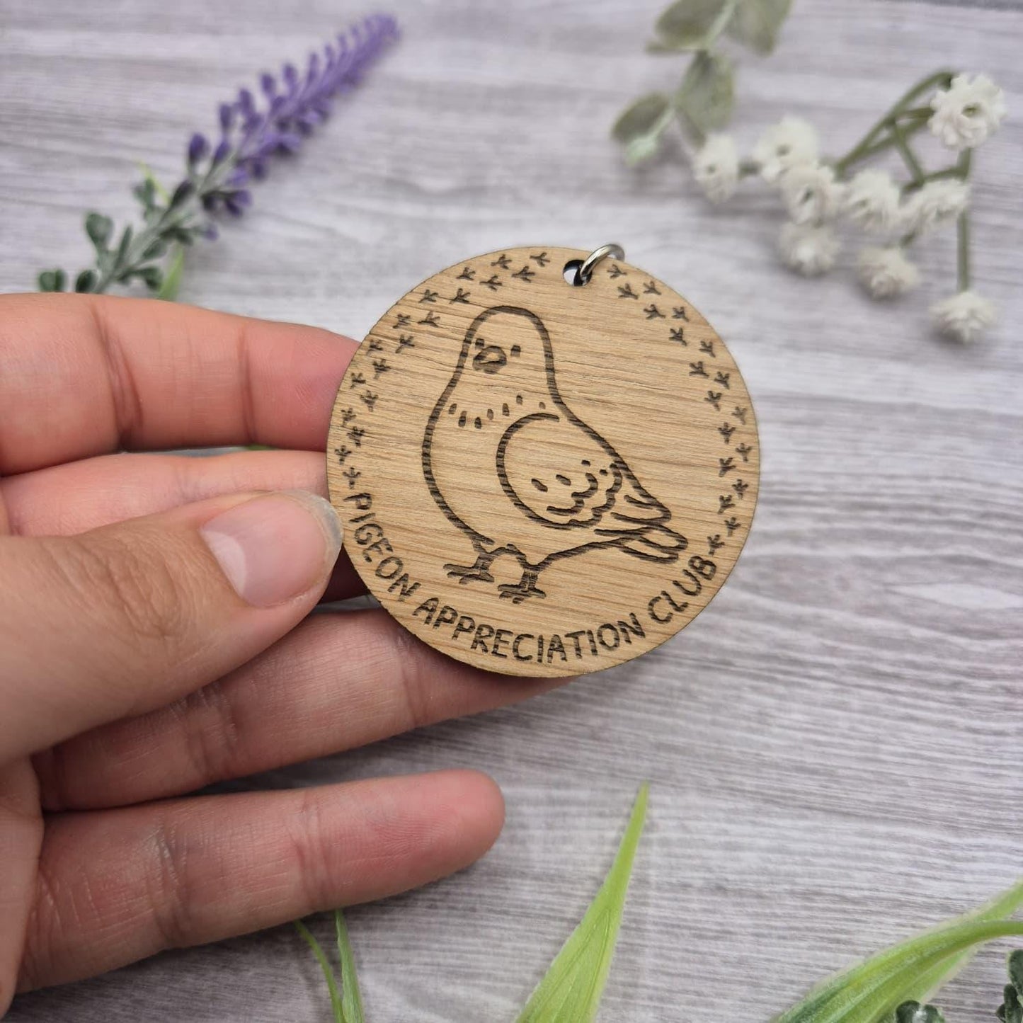 Wooden round keychain with pigeon illustration and 'Pigeon Appreciation Club' text, held by a hand on a wooden surface with flowers.