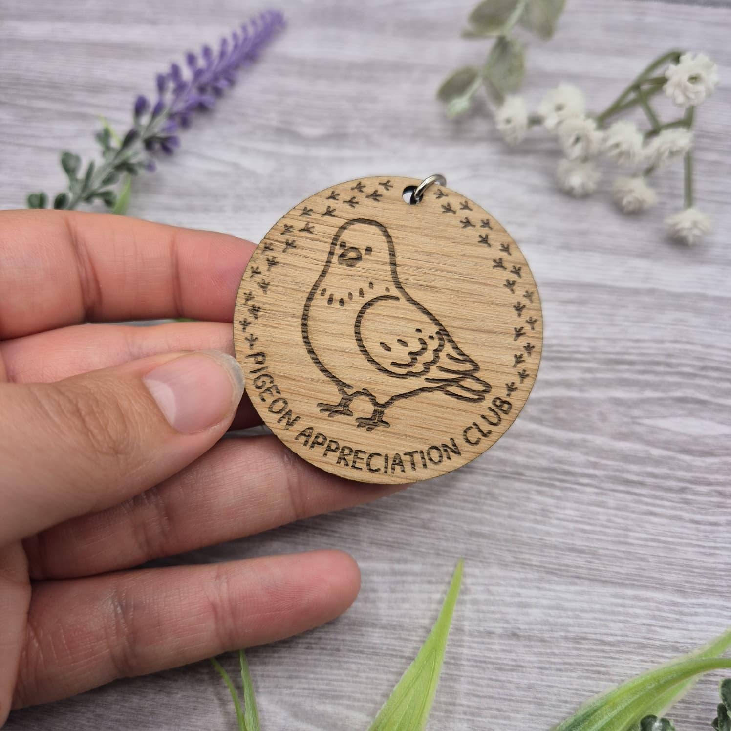 Wooden round keychain with pigeon illustration and 'Pigeon Appreciation Club' text, held by a hand on a wooden surface with flowers.