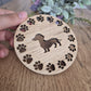A wooden round coaster with a sausage dog in the middle, with paw prints all around the outside.