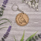 A wooden keychain with a bird engraved on it, surrounded by purple flowers and green leaves on a wooden surface.