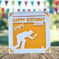 A birthday card with a silhouette of a person kneeling , playing crown green bowling and a banner with a star in the background, placed on a wooden surface.