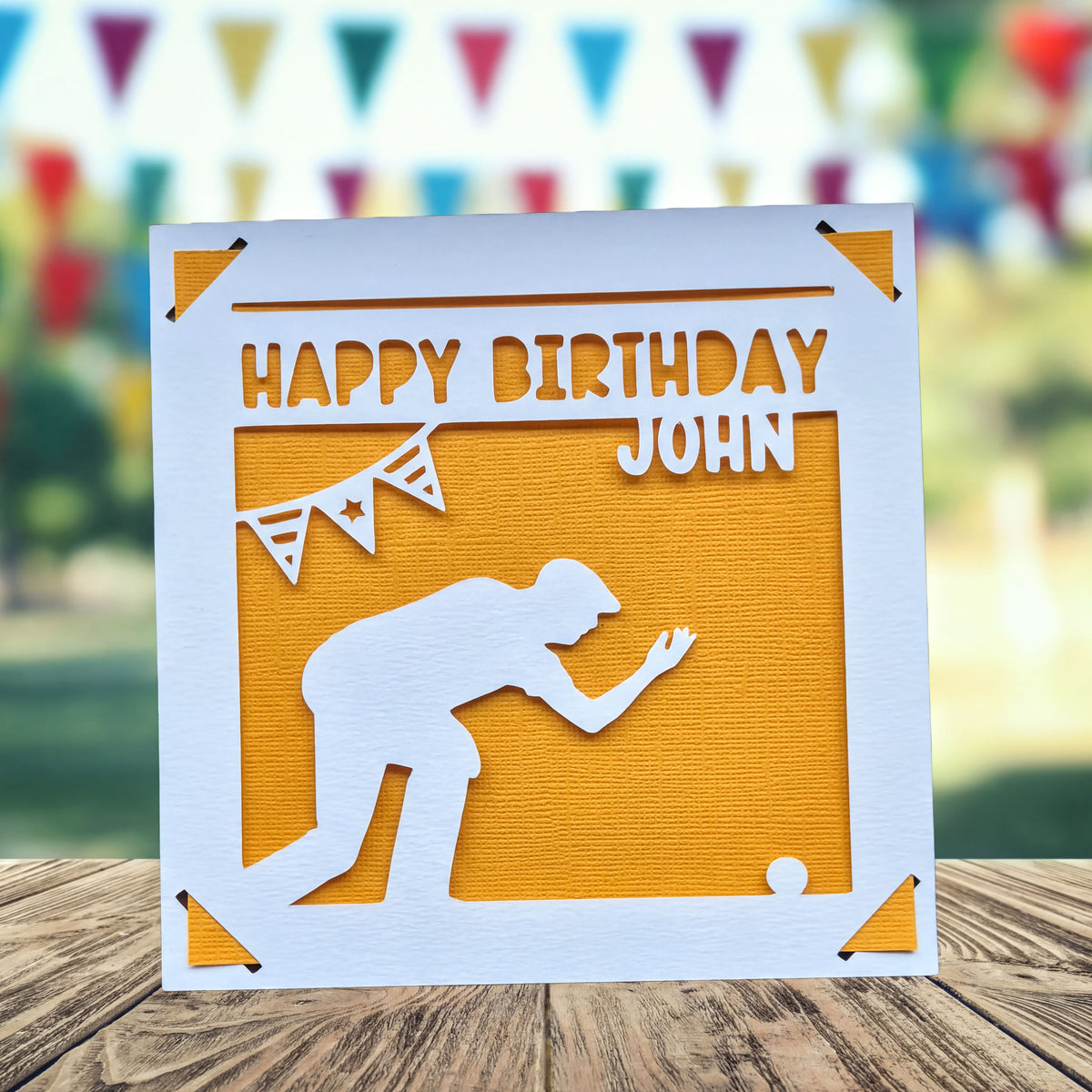 A birthday card with a silhouette of a person kneeling , playing crown green bowling and a banner with a star in the background, placed on a wooden surface.