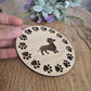 A wooden round coaster with a sausage dog in the middle, with paw prints all around the outside.