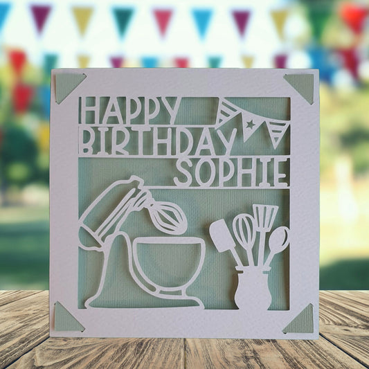 A personalised birthday card with a graphic of a baking mixing bowl and utensils, with the text 'Happy Birthday' and a space to add a name and age if required. The background is light green.