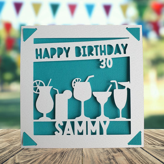 A personalized birthday card with a cocktail theme, featuring a white background with a cut-out silhouette of a cocktail glass and bottle, with the text 'Happy Birthday' and a space to add a name and age if required. The background colour is turquoise.