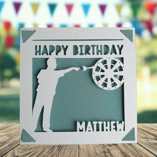A personalised birthday card with a darts theme, featuring a silhouette of a dart player on the front, with the text 'Happy Birthday' and a space to add a name and age if required.