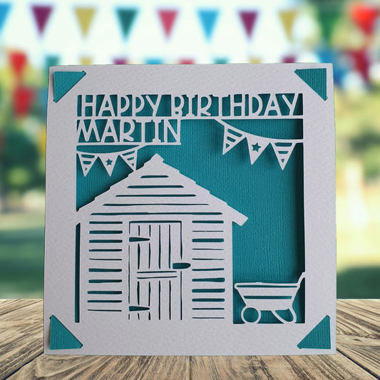 A personalised birthday card with a gardening shed illustration, made from hammered white card, with the text 'happy birthday' and the option to add a name and age. Card background is turquoise.