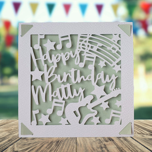 A personalised birthday card with a white guitar-shaped design, made from hammered white card, with the text 'Happy Birthday' and a space to add a name and age if required. Light green background