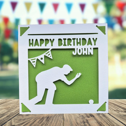 A white greeting card with a silhouette of a person kneeling, playing crown green bowling and a green background, along with the text "HAPPY BIRTHDAY JOHN" printed on it.