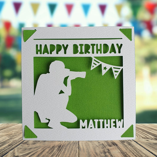 A personalised birthday card with a photographer theme, featuring a silhouette of a person with a camera, taking a picture, with the text 'Happy Birthday' and a space to add a name and age if required. Background is green.