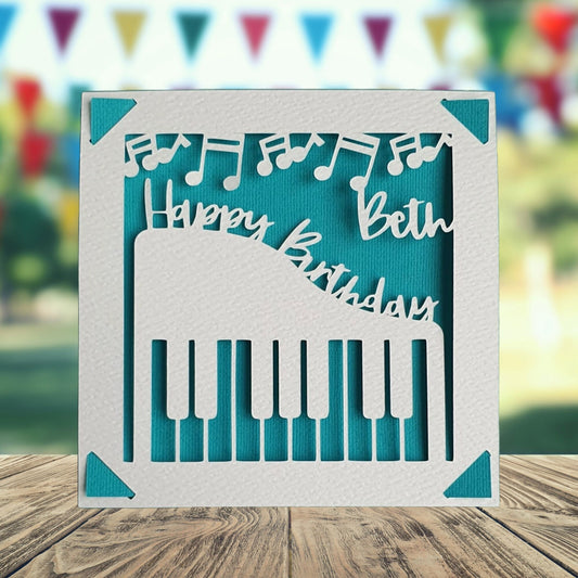 A personalised birthday card with a cut out of a piano on the front with musical notes at the top of the card, with the text 'Happy Birthday' and a space to add a name and age if required. The background is turquoise.