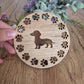 A wooden round coaster with a sausage dog in the middle, with paw prints all around the outside.