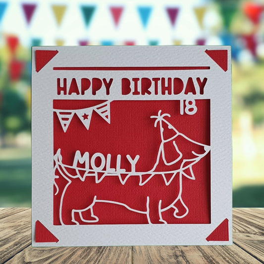 A personalised birthday card with a sausage dog illustration, made from white and red coloured card, with the text 'Happy Birthday' and a space to add a name and age if required.