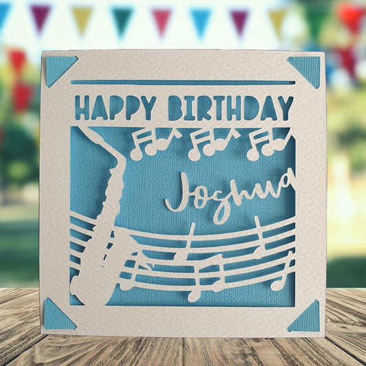 A personalised birthday card with a cut out of a saxophone on the front with musical notes, with a 'happy birthday' greeting and space to add a name and age. Card background is blue.